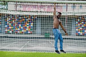 hübscher sexy afroamerikanischer muskulöser Mann mit nacktem Oberkörper in Jeans-Overalls posiert auf grünem Gras gegen Fußballtore des Stadionfeldes. modisches Porträt eines schwarzen Mannes. foto