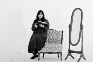 Attraktive südasiatische Frau in tiefrotem Kleid posiert im Studio auf weißem Hintergrund vor Spiegel und Stuhl, liest ein Buch. foto