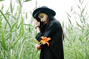 sinnliches Mädchen ganz in Schwarz, roten Lippen und Hut. gotische dramatische frau hält orangefarbene lilienblume auf gemeinem schilf. foto