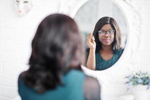 hübsche afroamerikanische frau mit brille, die im raum gegen den spiegel posiert. foto