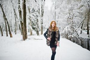 Rothaariges Mädchen im Pelzmantel, das im verschneiten Winterpark spazieren geht. foto
