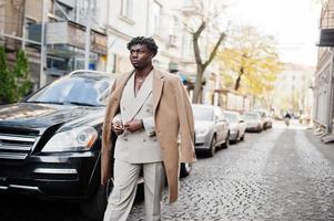 Stilvoller Afro-Mann in beigefarbenem Anzug und Mantel der alten Schule, der gegen ein schwarzes Geschäftsauto läuft. Modischer junger afrikanischer Mann in Freizeitjacke auf nacktem Oberkörper. foto