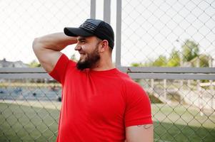 junger brutaler bärtiger muskulöser mann trägt rotes hemd, shorts und mütze im stadion. foto