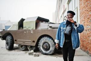 afroamerikanischer mann in jeansjacke, baskenmütze und brille, rauchende zigarre und posierte gegen btr militärisches gepanzertes fahrzeug. foto