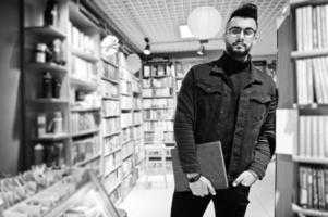 großer, intelligenter arabischer student, trägt schwarze jeansjacke und brille, in der bibliothek mit buch zur hand. foto