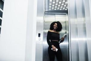 modische junge schöne afroamerikanische geschäftsfrau mit afro-frisur tragen in elegantem schwarz, stehen am aufzug mit telefon und laptop zur hand. foto