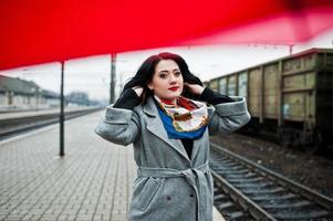 Brünettes Mädchen im grauen Mantel mit rotem Regenschirm im Bahnhof. foto