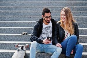 Cooles gemischtrassiges Paar, das mit Longboard auf der Treppe sitzt und ins Handy schaut. foto