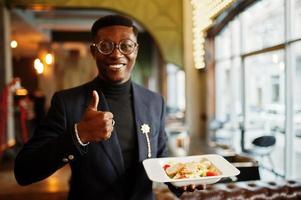Modischer afrikanisch-amerikanischer Mann in Anzug und Brille, der mit einem Teller Salat in der Hand im Café steht und den Daumen nach oben zeigt. foto