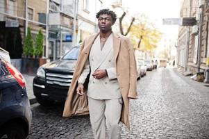 Stilvoller Afro-Mann in beigefarbenem Anzug und Mantel der alten Schule, der gegen ein schwarzes Geschäftsauto läuft. Modischer junger afrikanischer Mann in Freizeitjacke auf nacktem Oberkörper. foto
