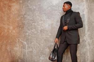 starker, mächtiger afroamerikanischer mann im schwarzen anzug und rollkragenpullover mit handtasche, die an der grauen wand posiert. foto