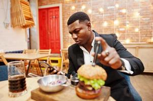 respektabler junger afroamerikanischer mann im schwarzen anzug, der im restaurant mit leckerem doppelburger und limonadengetränk sitzt. Blick aufs Telefon vor dem Essen. foto