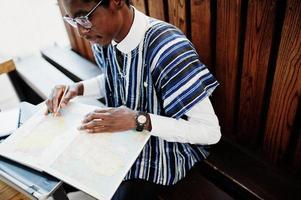 afrikanischer Mann in traditioneller Kleidung und Brille sitzt hinter Laptop im Café im Freien und schaut auf der Karte von Afrika und Ghana auf sein Notizbuch. foto