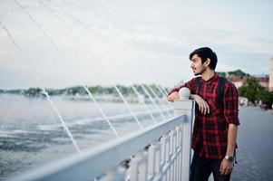 junger indischer studentenmann in kariertem hemd und jeans mit rucksack posierte in der abendstadt gegen brunnen. foto