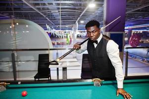 Junger gutaussehender afrikanischer Mann mit weißem Hemd, schwarzer Weste und Fliege spielt Billard. foto