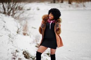 lockiges haar afroamerikanerin trägt schaffellmantel und handschuhe, die am wintertag gegen den zugefrorenen see gestellt werden. foto