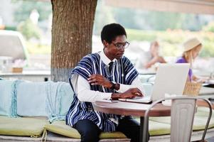 afrikanischer mann in traditioneller kleidung und brille sitzt hinter laptop im outdoor-café. foto