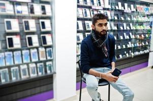 indischer bart mann kunde käufer im handyladen sitzt auf stuhl mit smartphone zur hand. Konzept der südasiatischen Völker und Technologien. handy shop. foto