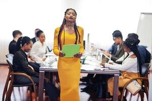 Gesicht einer hübschen afrikanischen Geschäftsfrau in gelbem Kleid, die eine Zwischenablage auf dem Hintergrund eines gemischtrassigen Teamtreffens der Geschäftsleute hält und am Bürotisch sitzt. foto