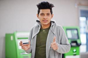 junger stilvoller asiatischer mann mit kreditkarte in der hand zeigt daumen hoch gegen reihe von grünen geldautomaten. foto