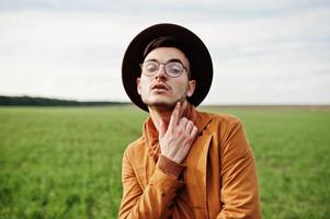 stylischer mann mit brille, brauner jacke und hut posierte auf der grünen wiese. foto