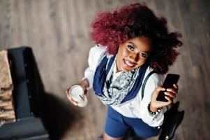 Attraktives afroamerikanisches lockiges Mädchen in weißer Bluse und blauen Shorts posierte im Café mit Latte und Handy zur Hand. Sicht von oben.