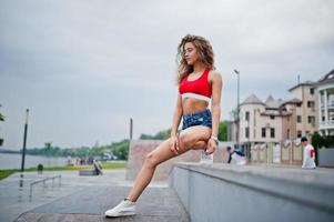 sexy lockiges Model-Mädchen in rotem Oberteil, Jeans-Denim-Shorts, Brille und Turnschuhen posiert im Skatepark. foto