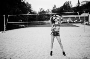 sexy lockiges Model-Mädchen in rotem Top, Jeans-Denim-Shorts, Tasse und Sonnenbrille am Beach-Volleyball-Feld. foto