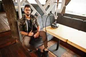 stylischer bart arabischer mann in brille und militärjacke raucht shisha an der straßenbar. Arabisches Model, das sich ausruht. foto