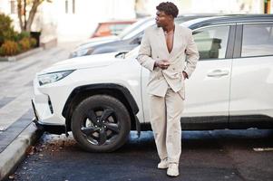 Stylischer Afro-Mann in beigem Old-School-Anzug gegen modernes Hybrid-Elektro-SUV-Auto. Modischer junger afrikanischer Mann in Freizeitjacke auf nacktem Oberkörper. foto
