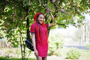 Süßes und schlankes afroamerikanisches Mädchen in rotem Kleid mit Dreadlocks posierte im Frühlingspark im Freien. stylisches schwarzes Modell. foto