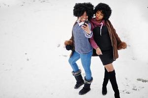 zwei lockige haare afroamerikanerinnen tragen einen schaffellmantel und handschuhe, die am wintertag gestellt wurden und selfie am telefon machen. foto