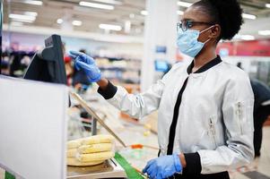 afrikanische frau mit medizinischer einwegmaske und handschuhen, die während des ausbruchs der coronavirus-pandemie im supermarkt einkaufen. schwarze frau wiegt früchte zur epidemiezeit. foto
