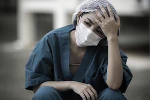 müde depressive asiatische krankenschwester trägt gesichtsmaske blaue uniform sitzt auf dem krankenhausboden, junge ärztin gestresst von harter arbeit foto