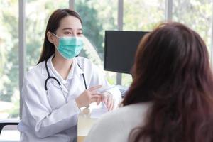 Die asiatische ältere Patientin wird von einer Ärztin auf Gesundheit untersucht, während beide in einem Untersuchungsraum eines Krankenhauses eine medizinische Gesichtsmaske tragen. foto