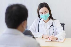 asiatische ärztin trägt eine medizinische gesichtsmaske und zeichnet symptome eines männlichen patienten auf papier im krankenhaus auf. Coronavirus-Schutzkonzept. foto