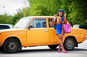 Schöne afrikanisch-amerikanische Dame mit Einkaufstüten, die in der Nähe eines orangefarbenen klassischen Retro-Autos steht. foto