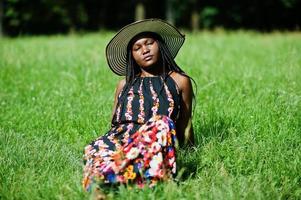 Porträt einer wunderschönen afroamerikanischen Frau der 20er Jahre mit Sommerhut, die auf grünem Gras im Park sitzt. foto