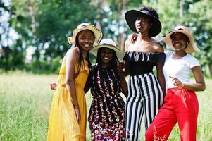 eine gruppe von vier wunderschönen afroamerikanischen frauen trägt einen sommerhut, der zeit auf grünem gras im park verbringt. foto