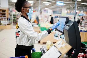 afrikanische frau mit medizinischer einwegmaske und handschuhen, die während des ausbruchs der coronavirus-pandemie im supermarkt einkaufen. schwarze frau wiegt früchte zur epidemiezeit. foto