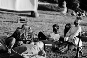 junge multiethnische gruppe von menschen, die filme im poof im open-air-kino ansehen, tragen während der covid-coronavirus-quarantäne eine maske. foto