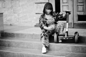 schöne afroamerikanische frau sitzt in der nähe von segway oder hoverboard. Schwarzes Mädchen mit selbstausgleichendem Elektroroller mit zwei Rädern und Handy. foto