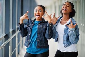 zwei afrikanische freundinnen in jeansjacke zeigen zusammen rockfinger drinnen. foto