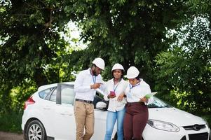 afroamerikanischer techniker in weißen helmen in der nähe des autos. gruppe von drei schwarzen ingenieuren treffen sich. foto