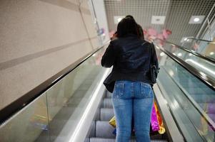 Rückseite einer afroamerikanischen Frau, die mit farbigen Einkaufstaschen im Einkaufszentrum auf der Rolltreppe spazieren geht. foto