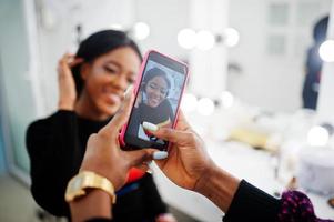 Afroamerikanerin beim Schminken durch Make-up-Künstlerin im Schönheitssalon. künstlerin macht foto auf handy von ihrer arbeit.