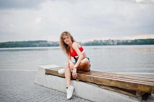 sexy lockiges Model-Mädchen in rotem Oberteil, Jeans-Denim-Shorts, Brille und Turnschuhen posiert auf einer Bank am See. foto