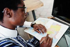 afrikanischer Mann in traditioneller Kleidung und Brille sitzt hinter Laptop im Café im Freien und schaut auf der Karte von Afrika und Ghana auf sein Notizbuch. foto