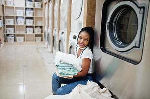 Fröhliche afroamerikanische Frau mit Handtüchern in der Nähe der Waschmaschine in der Selbstbedienungswäscherei. foto
