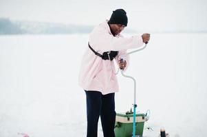 Afroamerikanischer Fischer, der mit Bohrer ein Loch in gefrorenes Eis bohrt. Winterangeln. foto